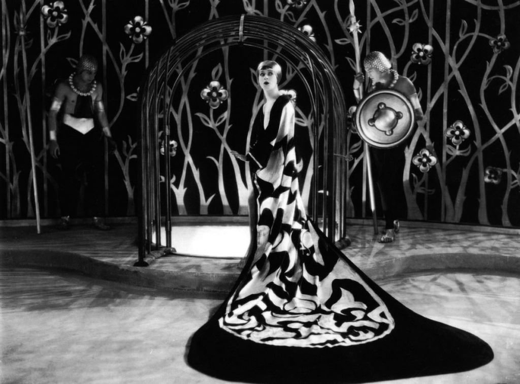 A black and white promotional still showing a woman in a long patterned dress with plunge neckline and large train looking at the camera. There id a lattice of what appears to be metal flowers behind her.