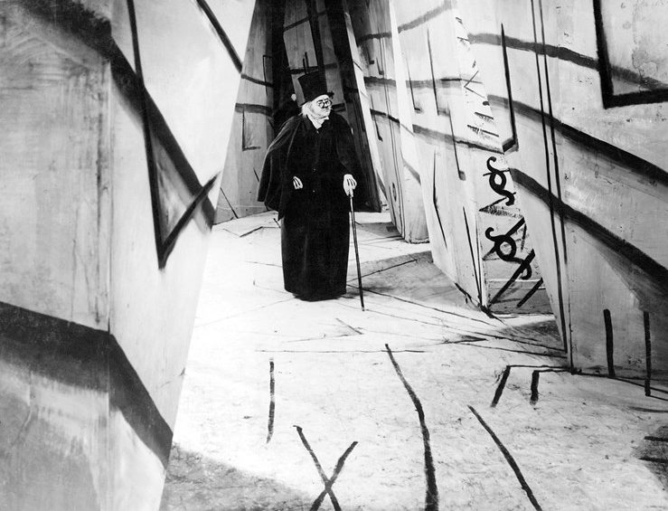 A black and white promotion still from the film, showing an old man with a cane and dressed all in black in a set surrounded by angular lines.